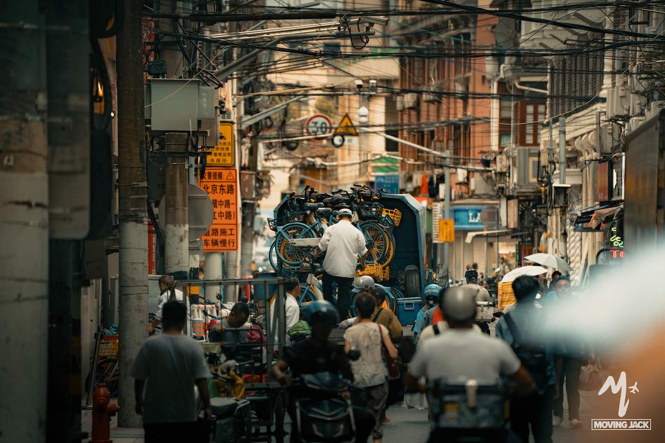 Een drukke stedelijke straatscène in Shanghai met mensen op fietsen en scooters, een arbeider die fietsen op een vrachtwagen laadt en bovengrondse elektriciteitskabels. Hoge gebouwen, borden met Chinese karakters en een bruisende energie verklaren waarom velen zich afvragen: is Shanghai een bezoek waard? -Copyright-moving-jack.com
