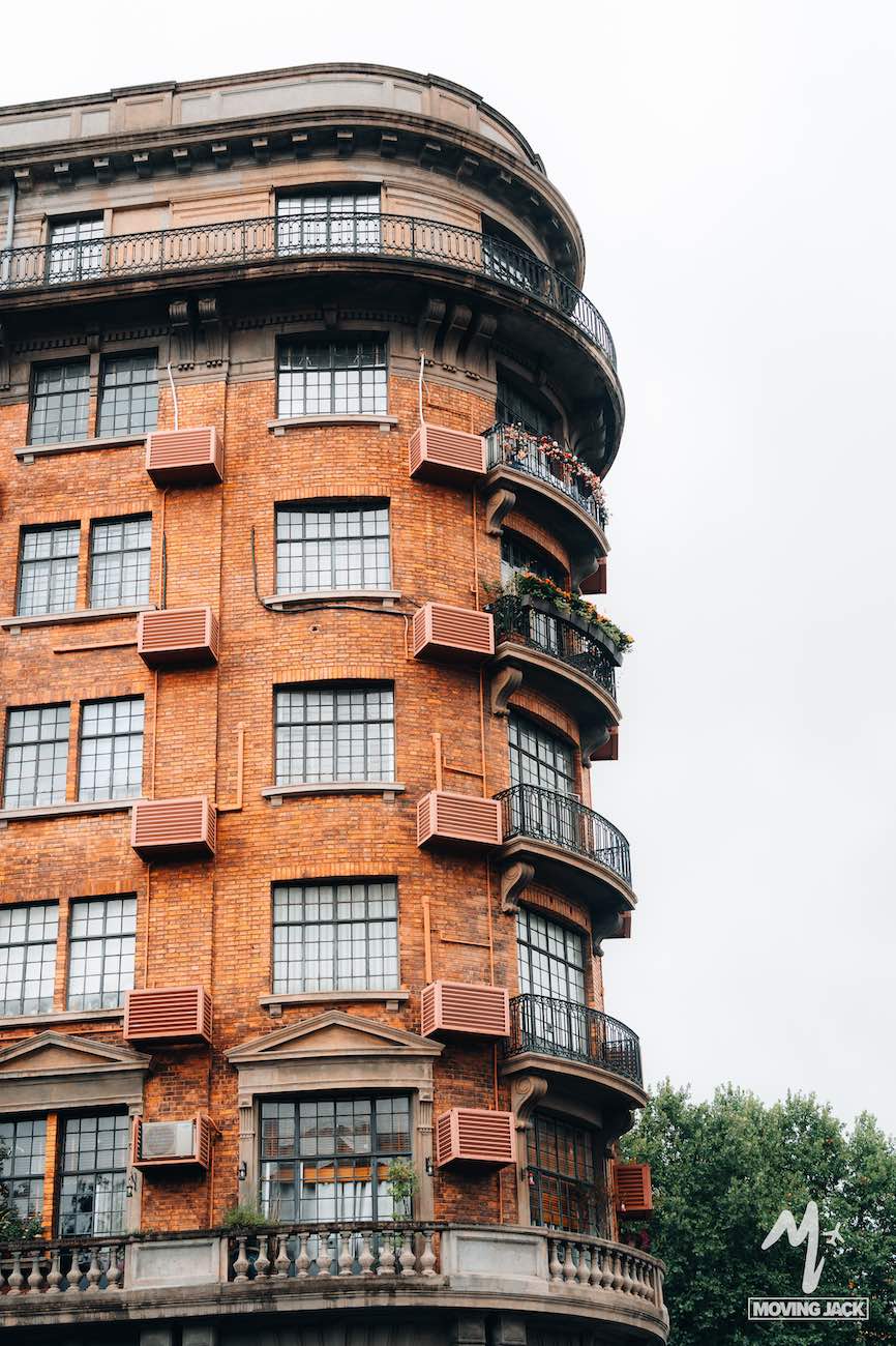 Een afgeronde hoek van een hoog, oranje bakstenen gebouw met grote ruitvormige ramen en roze luiken; groen en planten sieren het balkon – een alledaags tafereel dat je doet afvragen: is Shanghai de moeite waard om te bezoeken op een bewolkte dag? -Copyright-moving-jack.com