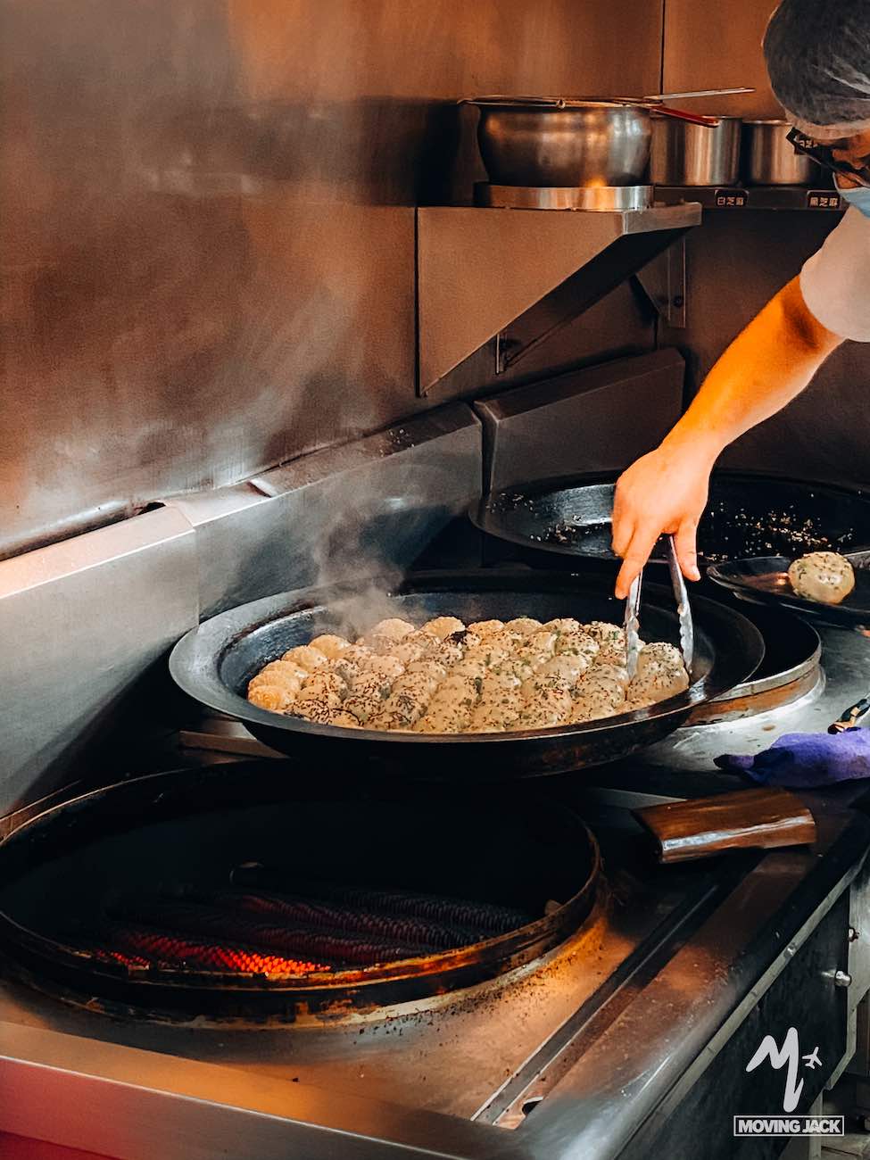 Iemand gebruikt een tang om een aantal gekruide gehaktballen te bakken in een grote zwarte pan op een fornuis, wat de vraag oproept: is Shanghai een bezoekje waard? Er stijgt stoom op uit de pan en er staan metalen bakjes op een plank erboven in deze professionele keuken. -Copyright-moving-jack.com