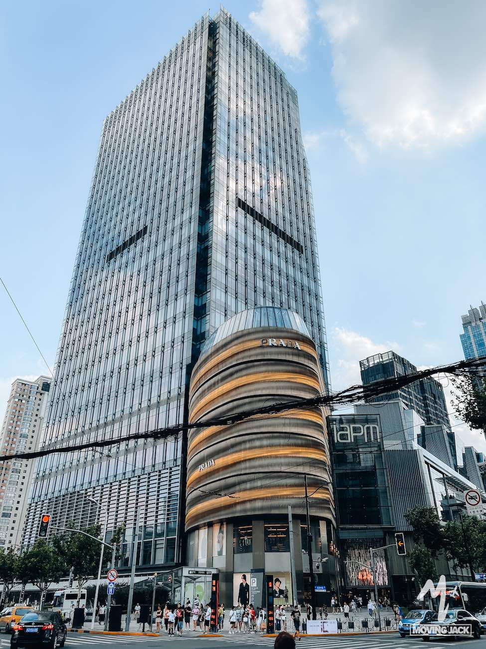 Een hoge glazen wolkenkrabber met een ronde, goudkleurige winkelpui aan de voet verrijst langs een drukke stadsstraat in Shanghai – een van de vele moderne gebouwen onder een gedeeltelijk bewolkte hemel, waardoor je je afvraagt: is Shanghai een bezoek waard? De levendige omgeving suggereert van wel. -Copyright-moving-jack.com