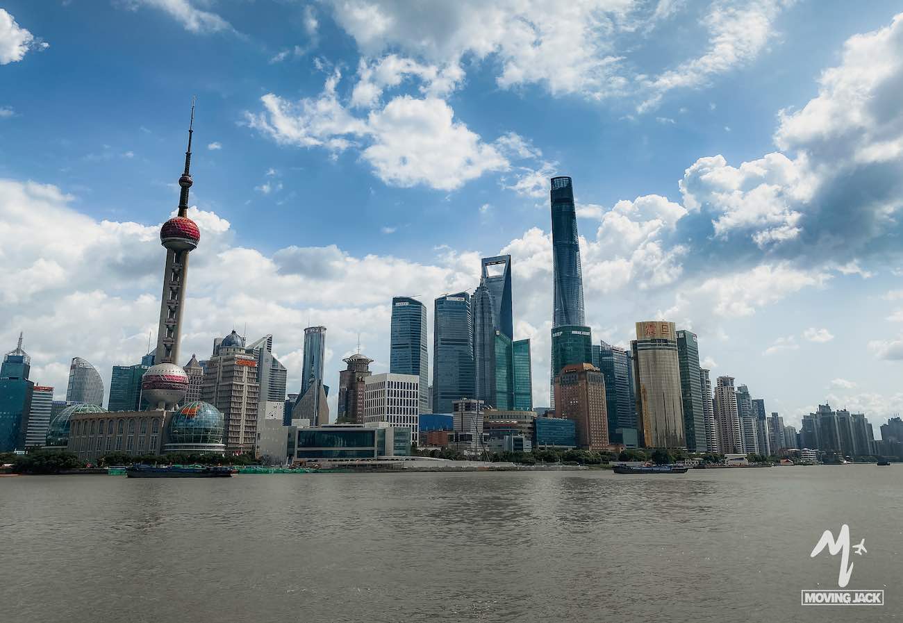 Shanghai skyline with modern skyscrapers, including the Oriental Pearl Tower, seen from across the Huangpu River under a partly cloudy blue sky—showcasing precisely why many wonder: Is Shanghai worth visiting?. -Copyright-moving-jack.com