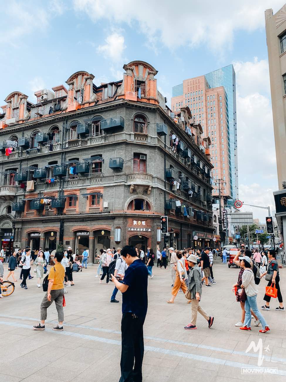 Een drukke stadsstraat toont veel mensen die voor een oud, meerlaags gebouw met balkons lopen, terwijl op de achtergrond moderne wolkenkrabbers oprijzen – wat de vraag oproept: is Shanghai de moeite waard om te bezoeken onder deze deels bewolkte hemel?.