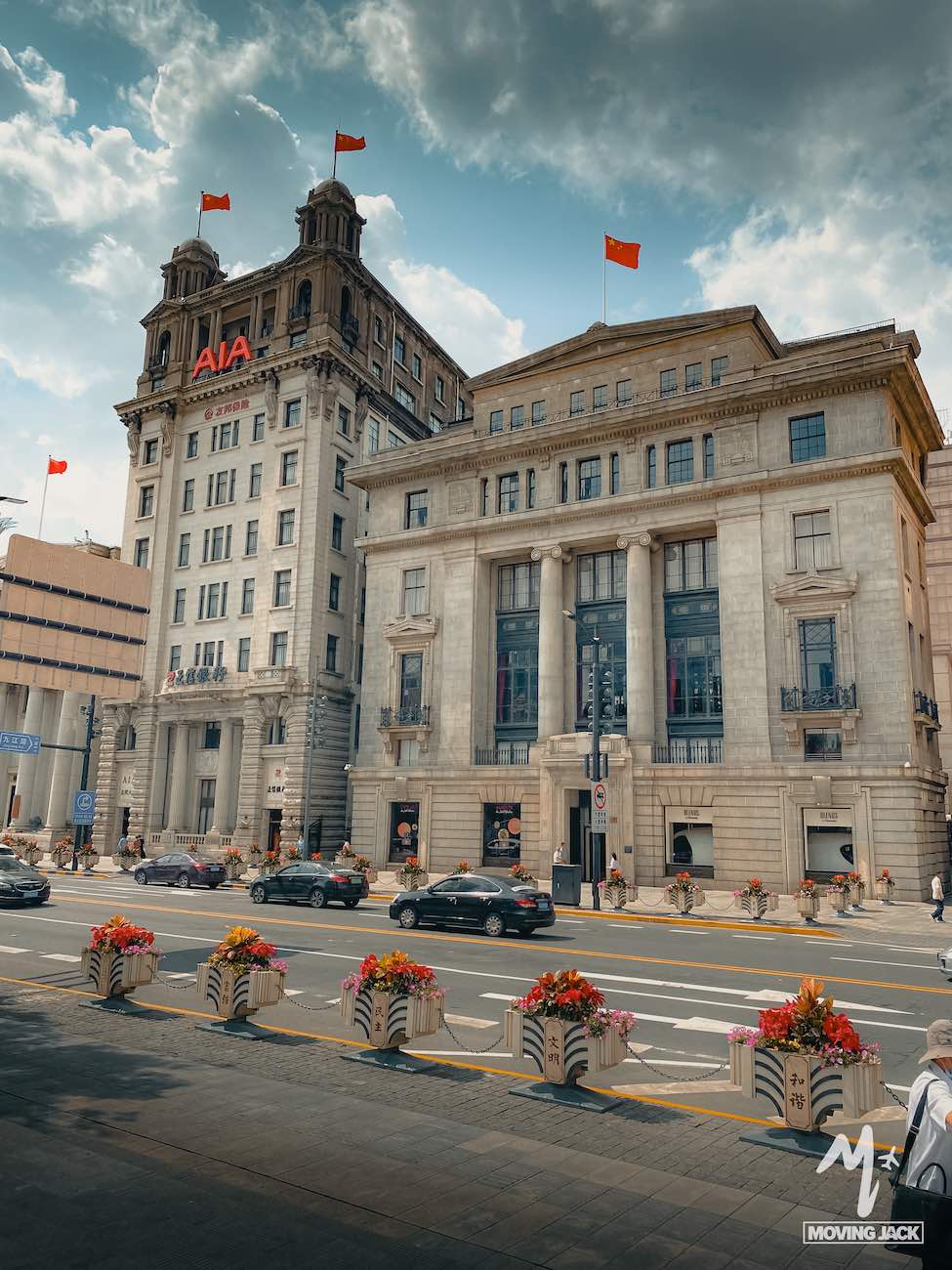 Een stadsstraat in Shanghai met historische gebouwen, waaronder een hoog gebouw met "AIA"-borden en rode vlaggen. Verschillende auto's rijden voorbij, kleurrijke bloembakken sieren de stoep en een gedeeltelijk bewolkte hemel geeft een hint naar de vraag die velen zich stellen: is Shanghai een bezoek waard?.