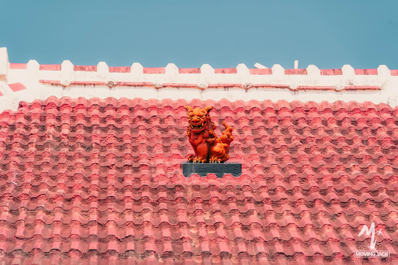 Een rood pannendak met in het midden een klein, feloranje Chinees leeuwenbeeldje (Foo Dog) tegen een helderblauwe hemel – een klassiek beeld dat je zeker moet zien op Okinawa. Witte sierlijsten sieren de rand van het dak. -Copyright-moving-jack.com