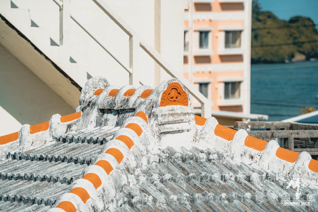 Een close-up van een traditioneel Chinees dak met witte en oranje decoratieve dakpannen, met op de achtergrond een trap en een pastelroze gebouw vlakbij het water – een unieke architectonische bezienswaardigheid te midden van alle activiteiten op Okinawa. -Copyright-moving-jack.com