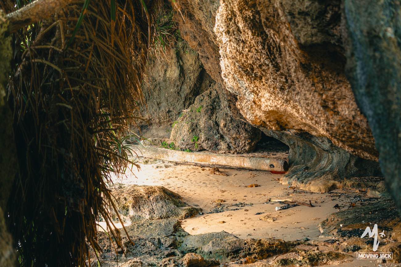 Een blik op een kleine zandgrot, omgeven door grote rotsen en hangende planten, met zonlicht dat door een opening schijnt – een van de verborgen pareltjes onder de bezienswaardigheden op Okinawa. Het Moving Mick-logo is zichtbaar in de rechterbenedenhoek. -Copyright-moving-jack.com