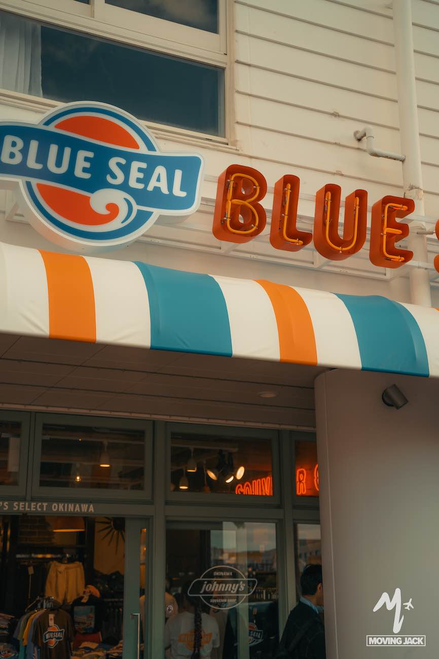 De afbeelding toont de buitenkant van een winkel genaamd "Blue Seal", met een kleurrijke blauw-oranje-witte gestreepte luifel en een fel neonbord boven de ingang – een van de iconische bezienswaardigheden in Okinawa. Mensen zijn zichtbaar bij de ingang. -Copyright-moving-jack.com