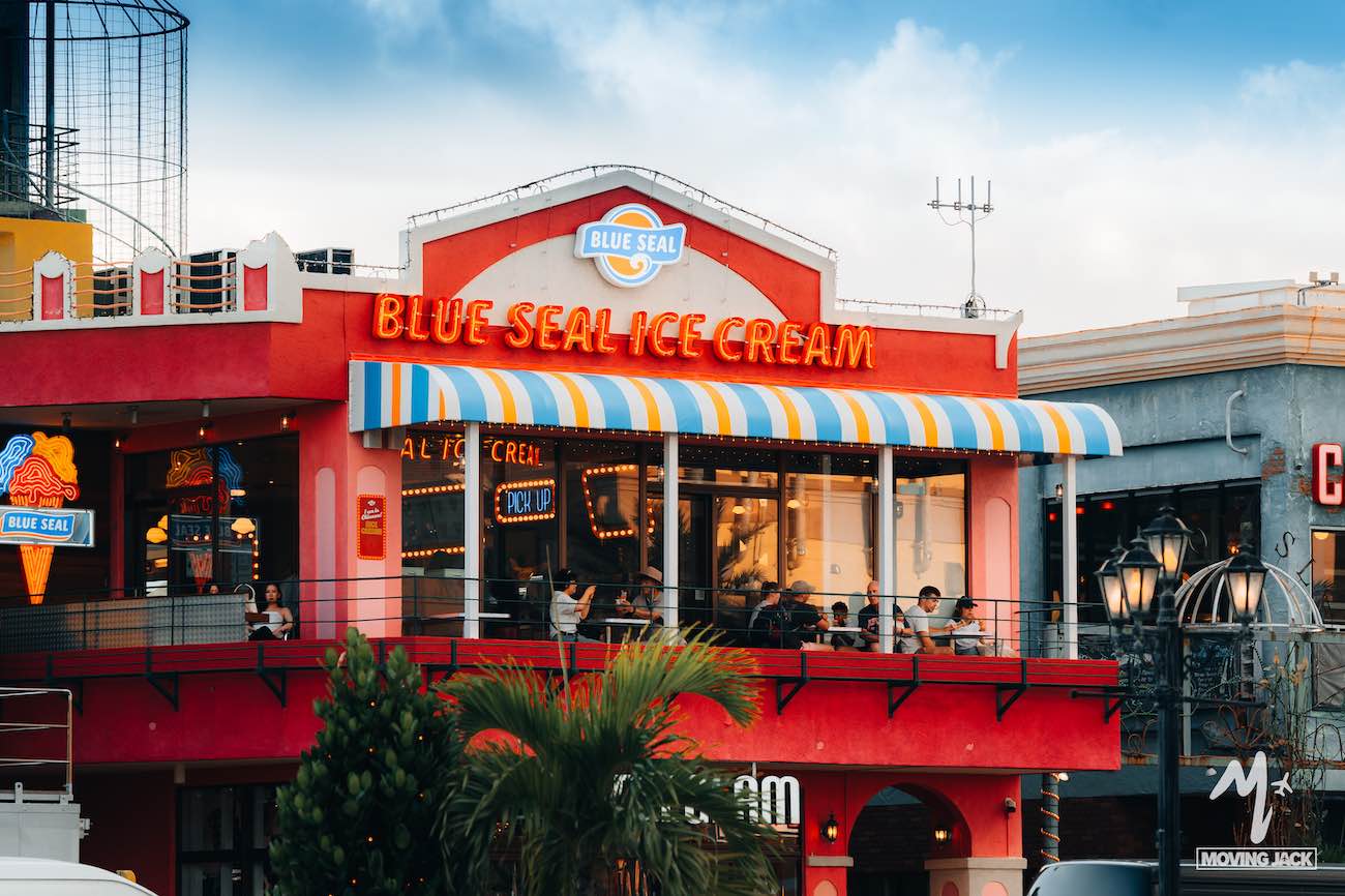 Een kleurrijke, twee verdiepingen tellende ijssalon met een Blue Seal-keurmerk, een gestreepte luifel en een terras, waar mensen genieten van een ijsje – een absolute aanrader tijdens een bezoek aan Okinawa. Het gebouw is uitgevoerd in felrode en oranje tinten onder een gedeeltelijk bewolkte hemel. -Copyright-moving-jack.com