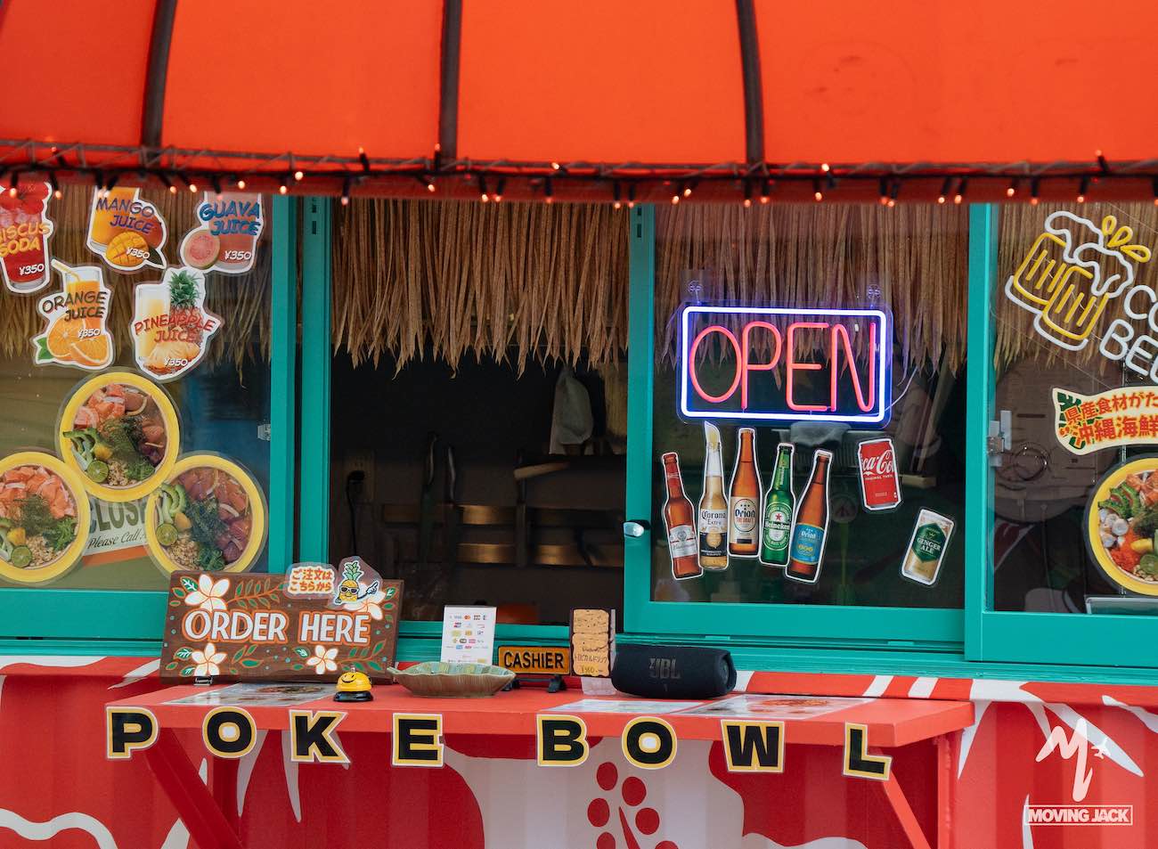 Een kleurrijke poké bowl-kraam met een neonbord met "OPEN", flesjes drank, foto's van het menu en een felrode luifel – een van de leuke dingen om te doen in Okinawa. Borden met "Bestel hier" en "POKÉ BOWL" zijn aanwezig, en tropische decoraties dragen bij aan de sfeer. -Copyright-moving-jack.com
