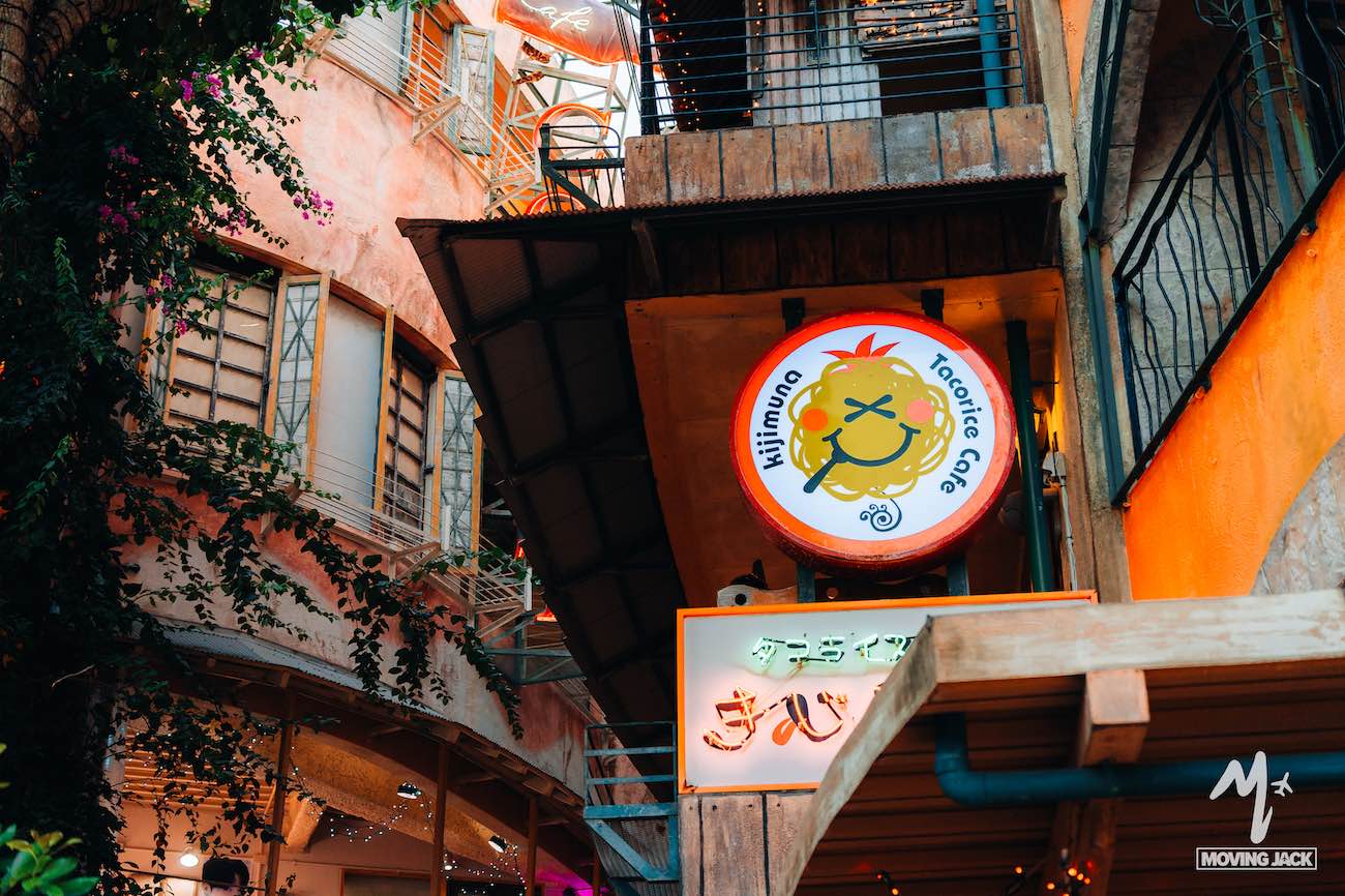 Een kleurrijk buitentafereel toont het uithangbord van "Mifimuno Tracutie Café" met een schattig berenlogo, verscholen tussen eclectische gebouwen en planten – een van de charmante dingen om te doen in de knusse steegjes van Okinawa. -Copyright-moving-jack.com