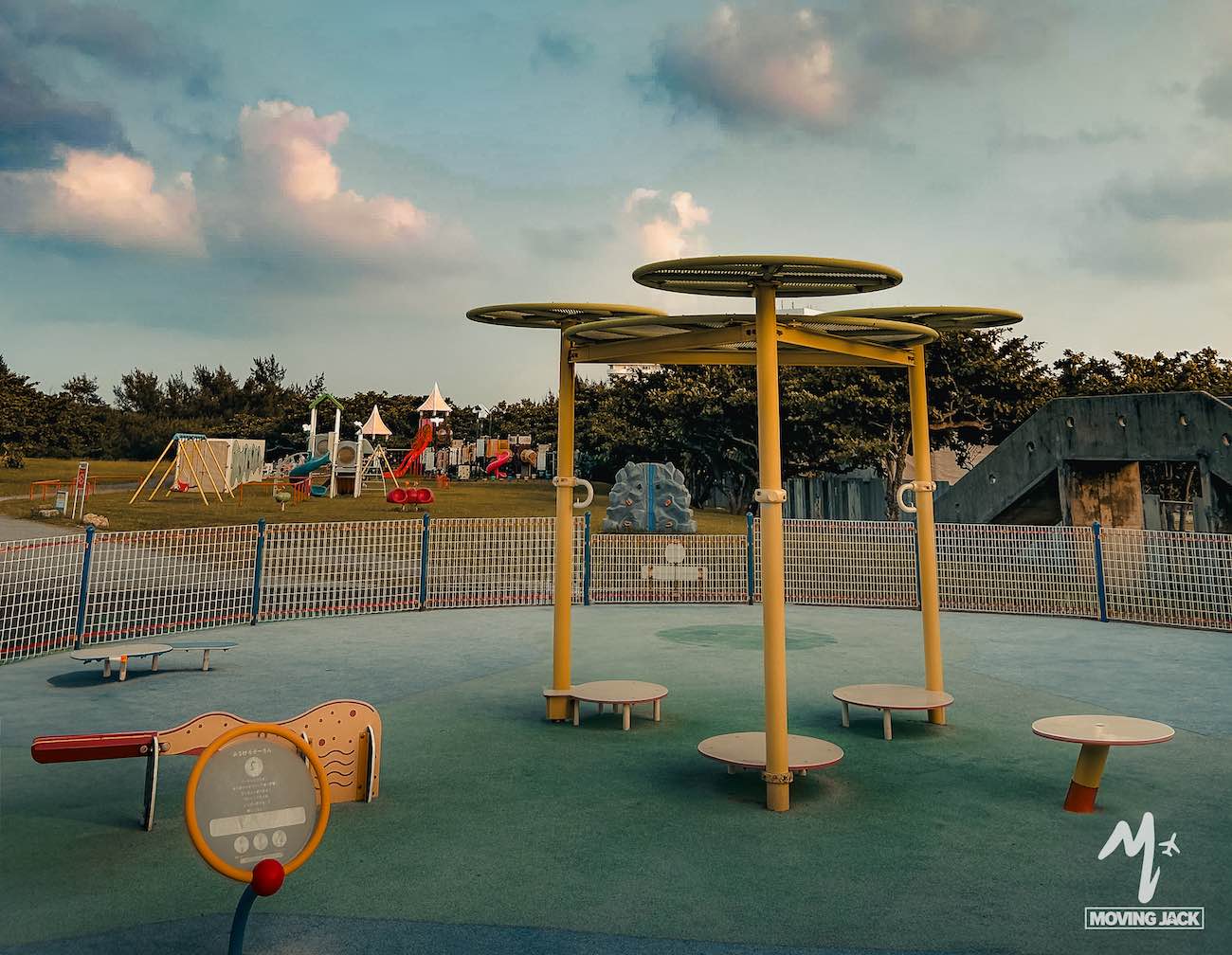 Een kleurrijke speeltuin, perfect voor gezinnen die op zoek zijn naar leuke activiteiten in Okinawa, met draaimolens op de voorgrond en glijbanen, klimtoestellen en groen gras op de achtergrond onder een gedeeltelijk bewolkte hemel. -Copyright-moving-jack.com