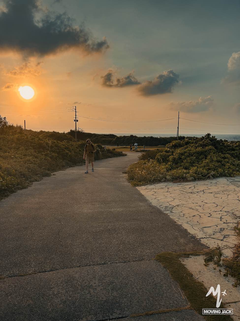 Een persoon wandelt alleen over een geplaveid pad bij zonsondergang, omgeven door groen – een van de rustgevende dingen om te doen in Okinawa. De zon staat laag en werpt warm licht en lange schaduwen onder een gedeeltelijk bewolkte hemel, wat een serene sfeer creëert. -Copyright-moving-jack.com