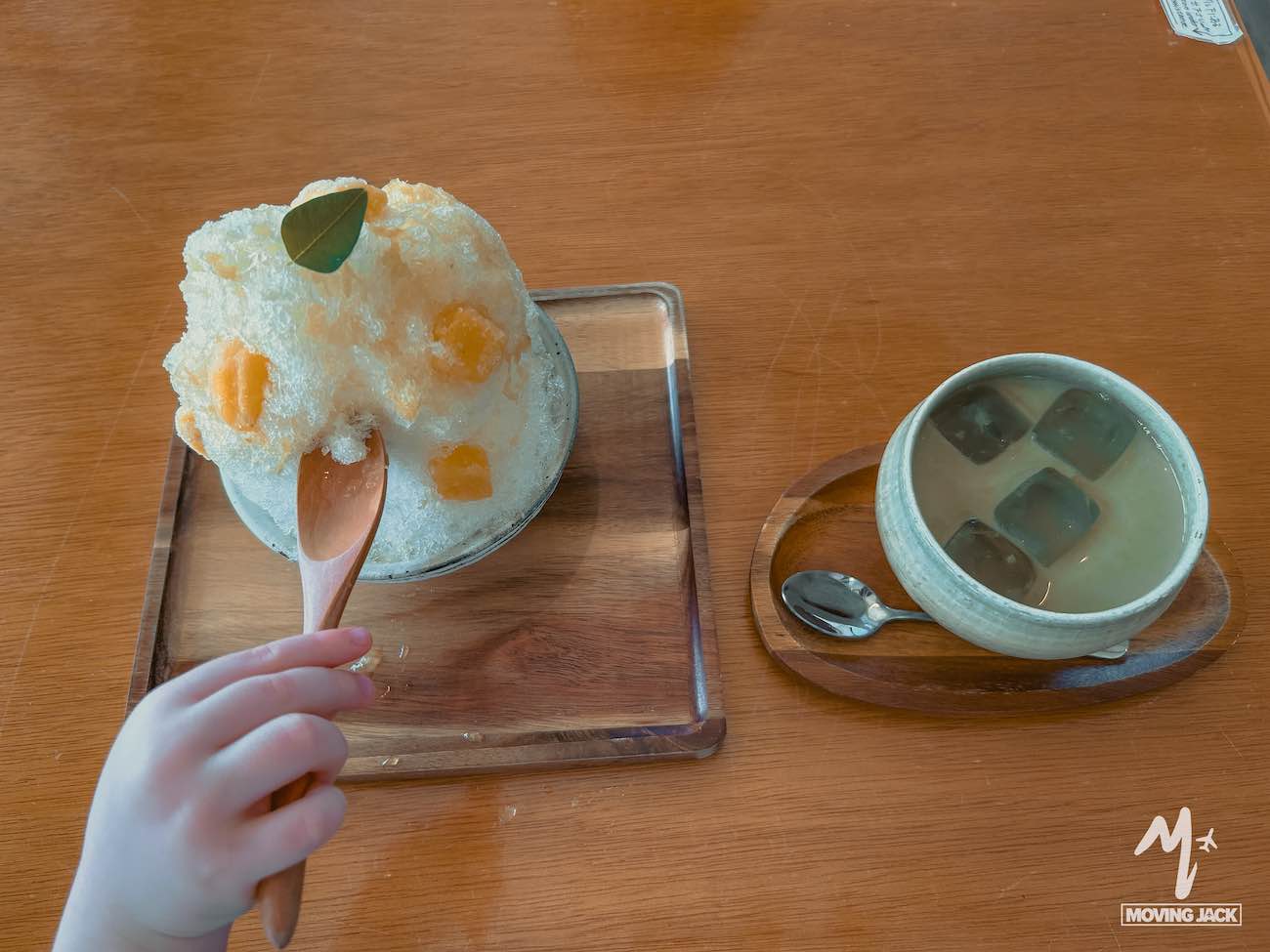 Een kinderhandje houdt een lepel vast en schept uit een kom met geschoren ijs en stukjes fruit – een van de heerlijke dingen om te doen in Okinawa – naast een kom met geleiblokjes in een melkachtige vloeistof op een houten tafel. -Copyright-moving-jack.com