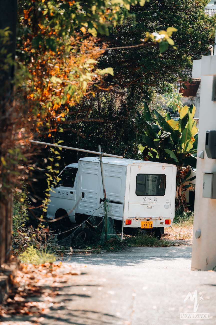 Een klein wit busje met Japanse opschriften op de zijkant staat geparkeerd aan een smal, zonnig pad, gedeeltelijk omgeven door groen en bomen – slechts één van de vele bezienswaardigheden die je kunt tegenkomen tijdens je ontdekkingstocht door Okinawa. Het voertuig lijkt enigszins scheef te staan tussen de planten. -Copyright-moving-jack.com