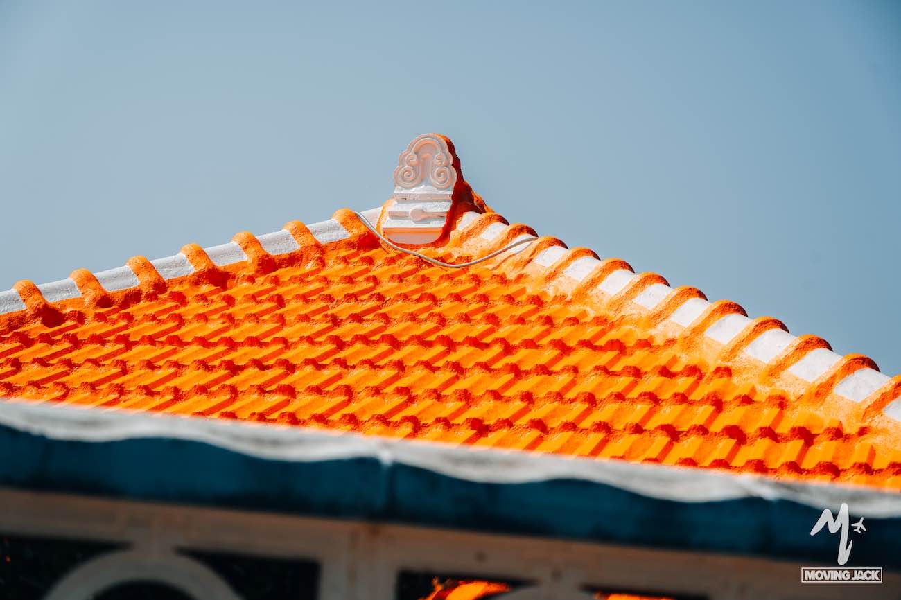 Een feloranje pannendak met witte nokken en een decoratief ornament op de top, afstekend tegen een helderblauwe hemel – een perfect plaatje om te bewonderen tijdens de vele activiteiten die Okinawa te bieden heeft. Een deel van een stenen muur is aan de onderkant zichtbaar. -Copyright-moving-jack.com