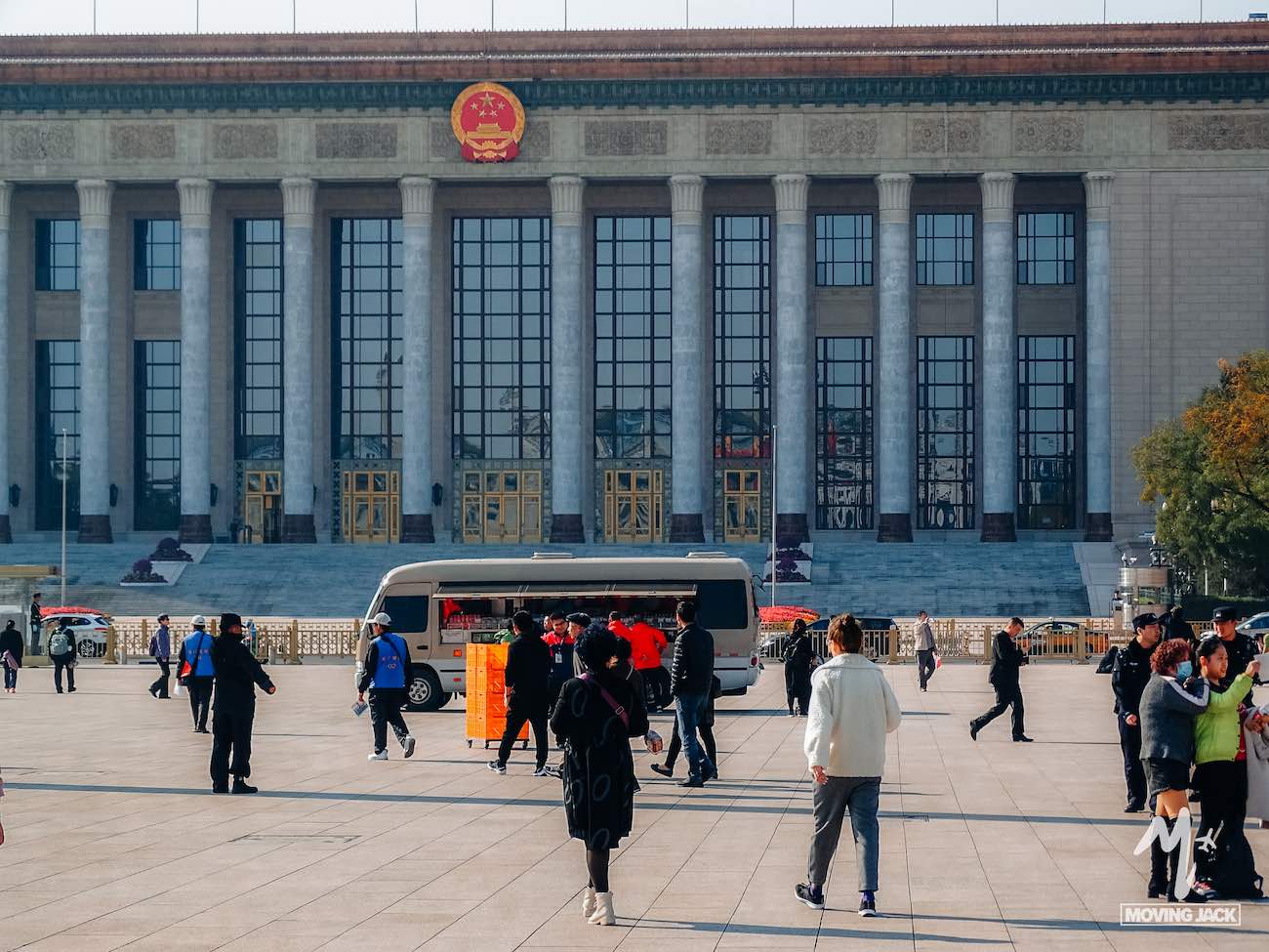 People walking and gathering in front of the Great Hall of the People, a landmark near popular options for where to stay in Beijing, with impressive columns and the national emblem of China displayed above the entrance.