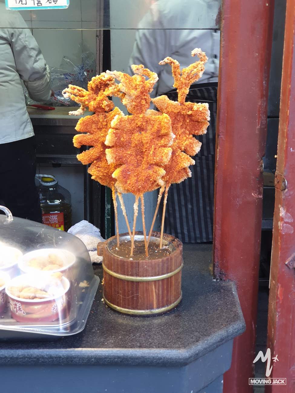 Several skewers of crispy, deep-fried food coated in orange crumbs are displayed upright in a wooden holder on a countertop near a street food stall—a must-try if you're exploring where to stay in Beijing.