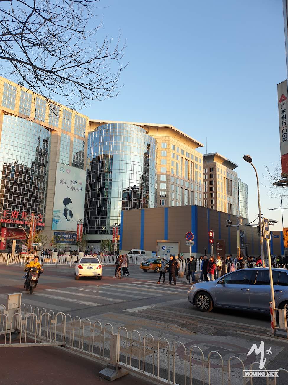 A city street scene with cars, pedestrians, and a large modern glass building reflecting sunlight in the background captures the lively energy of those exploring where to stay in Beijing.