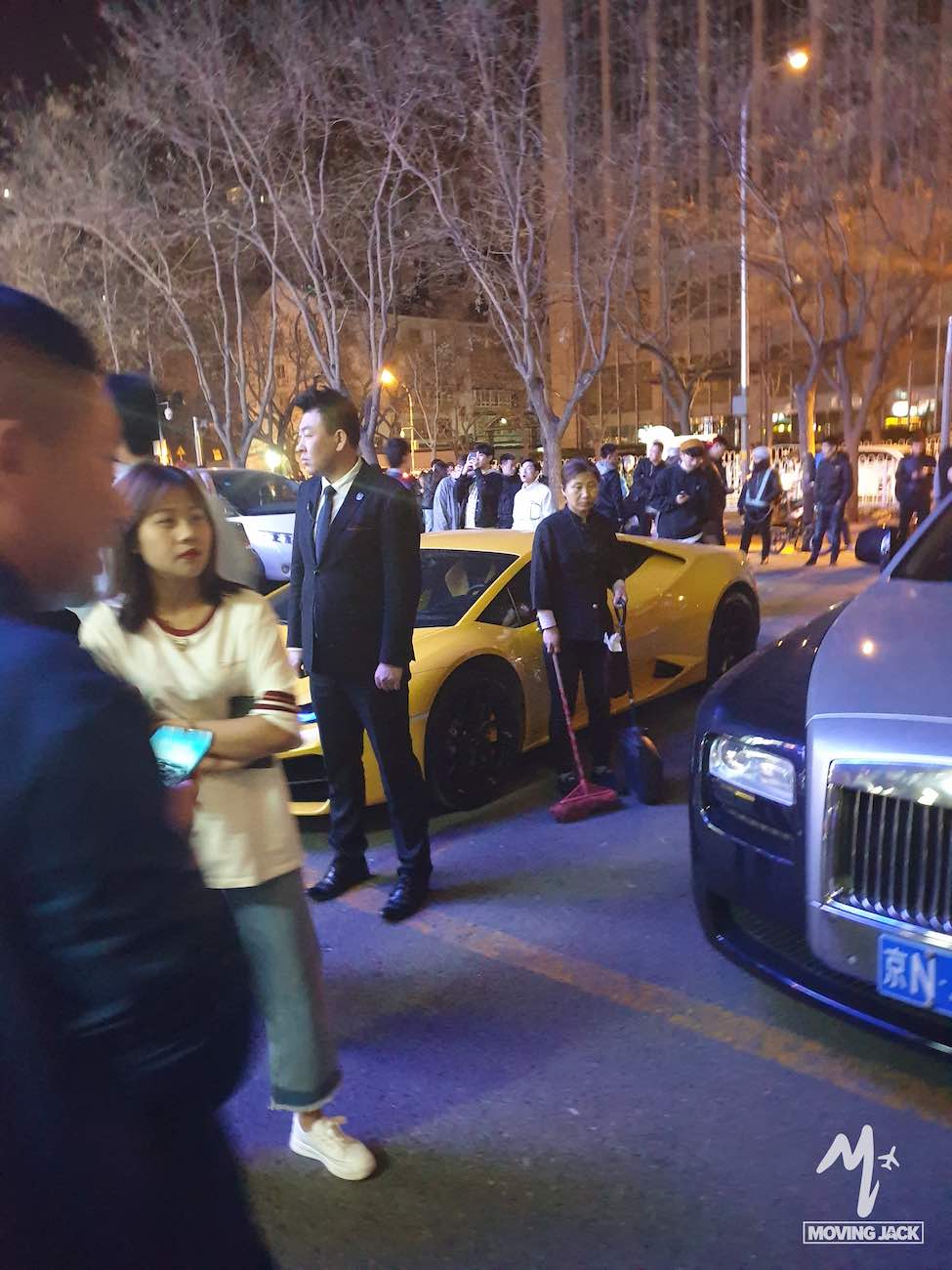 A crowd stands near luxury cars, including a yellow sports car and a gray car, on a city street at night. 