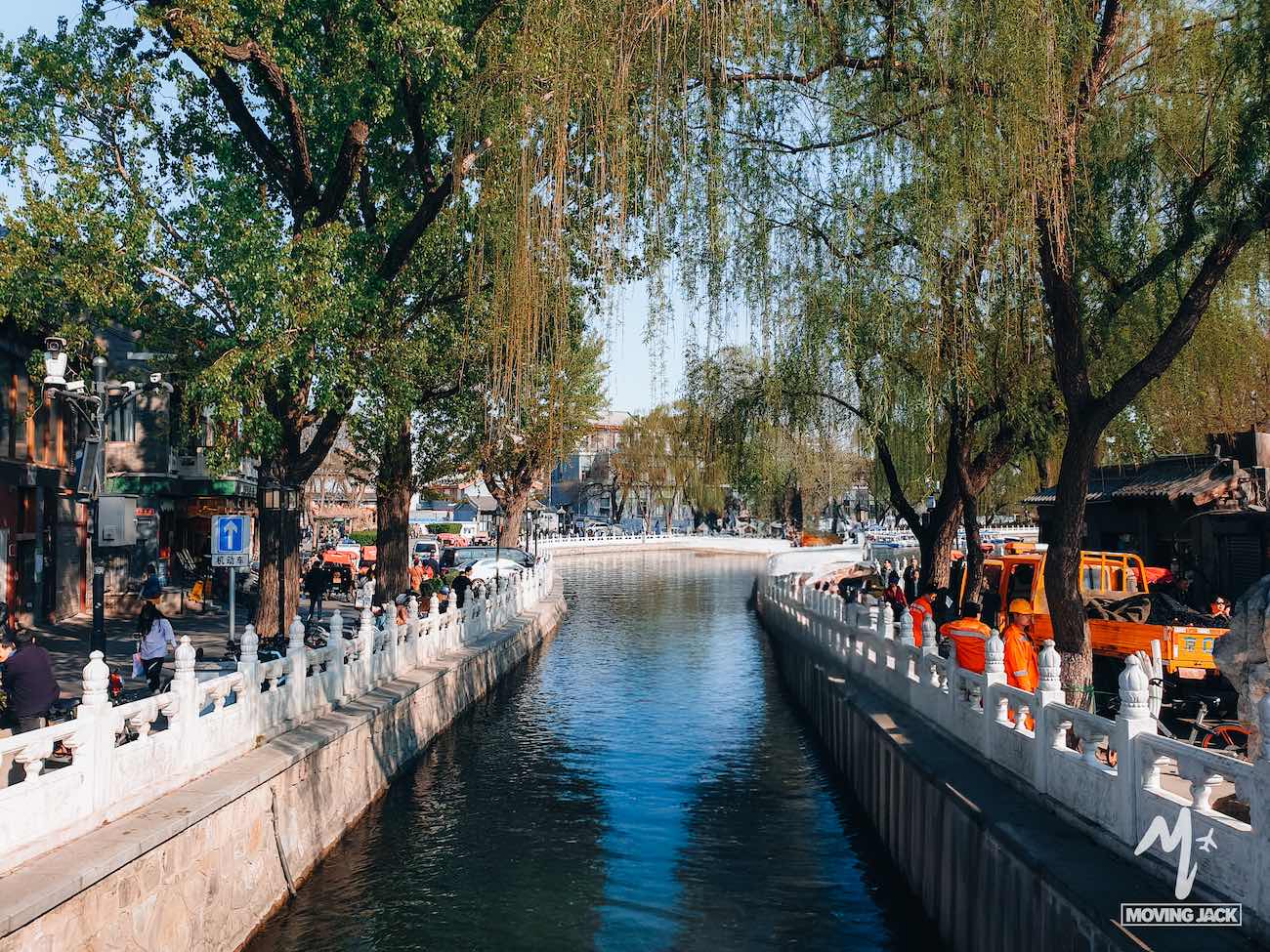 A canal lined with white railings and trees runs through an urban area, where people stroll along both sides—perfect for those exploring where to stay in Beijing—and maintenance workers in orange uniforms tend to the scene near orange vehicles.
