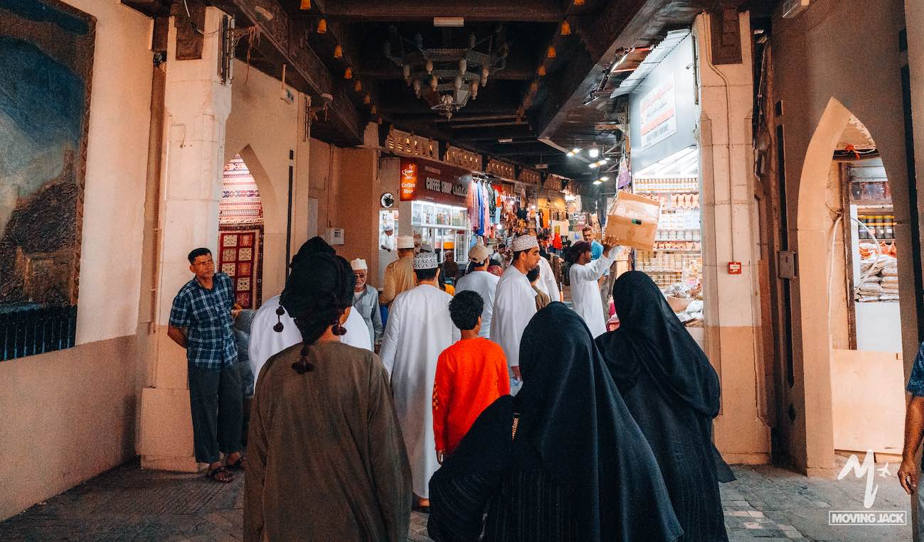 Mensen lopen door een bruisende overdekte markt met winkels aan weerszijden, sommigen gekleed in traditionele Midden-Oosterse kleding. De markt is druk, met kleurrijke waren en lichtjes op de achtergrond. -Copyright-moving-jack.com