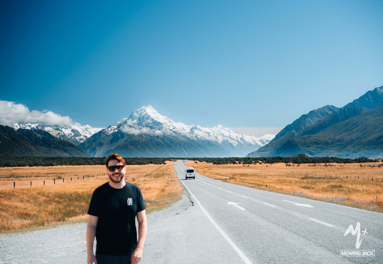 Smiling man stands on a road with mountains and a campervan in the background under a clear blue sky, capturing the spirit of exploring New Zealand by campervan—proving it's definitely worth it.