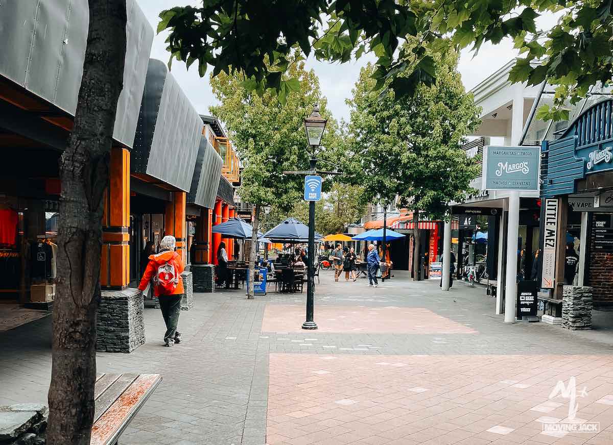 Outdoor shopping street with trees, shops, a few people walking, and outdoor seating with blue umbrellas—almost as inviting as exploring New Zealand by campervan, where every stop promises unique local charm.