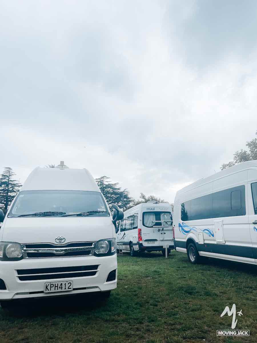 Several white vans and minibuses parked on grass under a cloudy sky, with trees in the background—a scene that makes you wonder, is New Zealand by campervan worth it?