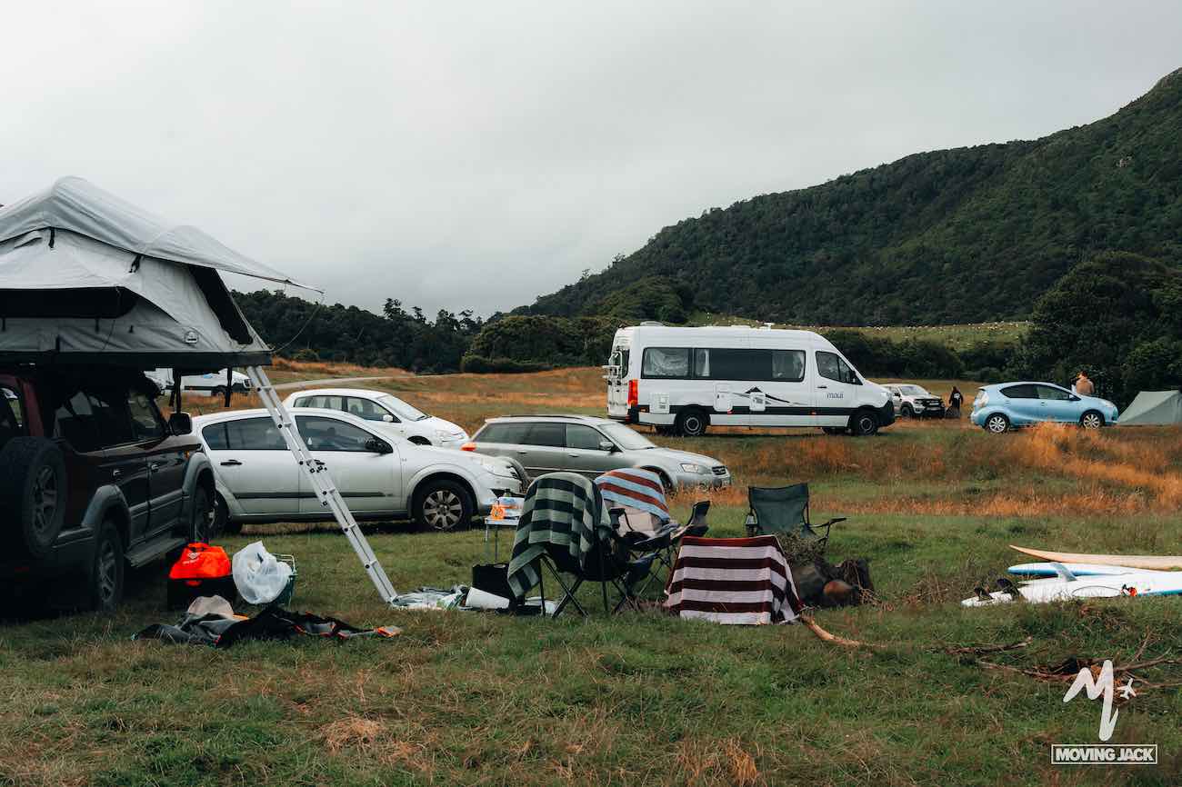 Camping site with chairs, vehicles, and tents set up on a grassy field near wooded hills under a cloudy sky—making you wonder, is New Zealand by campervan worth it? This spot perfectly captures the adventure and freedom of exploring by road.