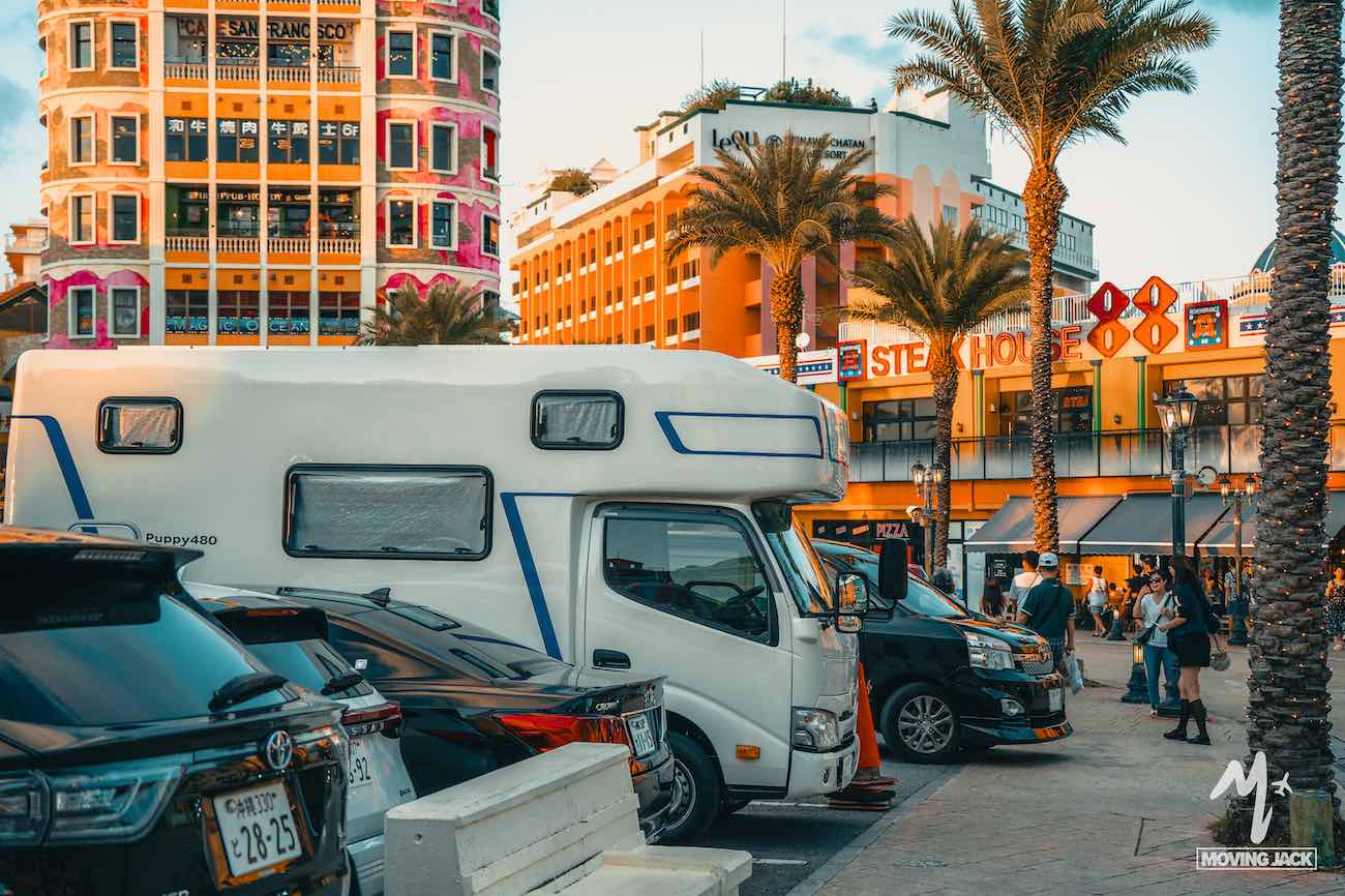 Een drukke straat met geparkeerde auto's en een camper, omgeven door kleurrijke gebouwen en palmbomen, geeft een beeld van het dagelijks leven in Okinawa – een levendige bestemming waar bezoekers vaak vragen: "Is Okinawa wel veilig?" De levendige scène weerspiegelt de gastvrije sfeer.