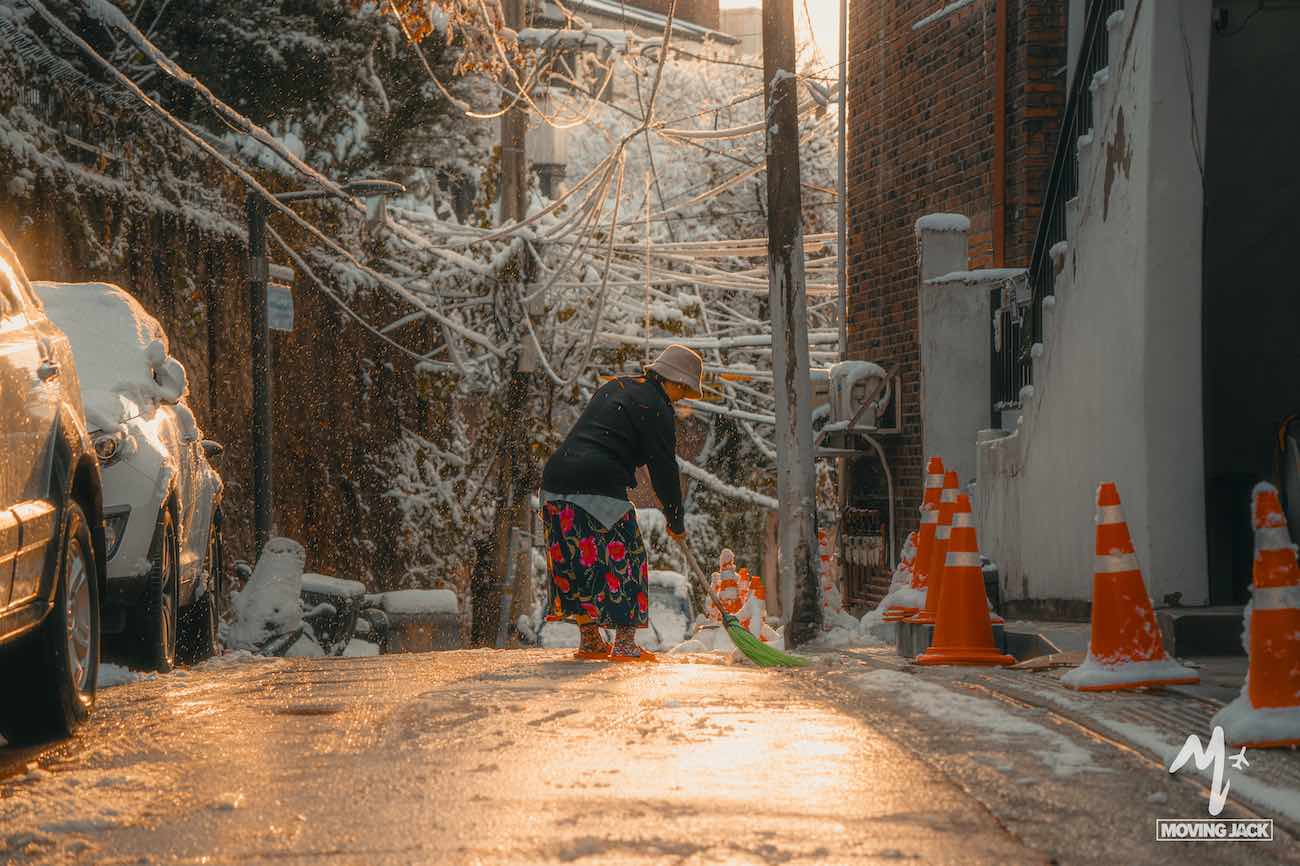 Een persoon schept sneeuw op een smalle, zonovergoten straat in Seoul – een perfecte plek voor je reisroute door Seoul – met oranje kegels en besneeuwde auto's en bomen.