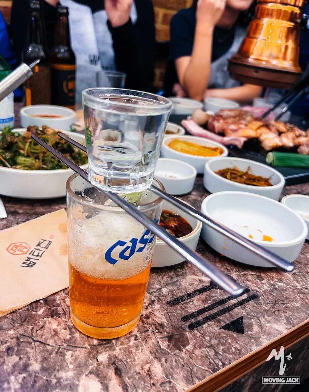 Een shotglaasje soju, balancerend op eetstokjes boven een glas bier tijdens een Koreaanse barbecue, met diverse bijgerechten – een absolute aanrader voor iedereen die Seoul bezoekt.