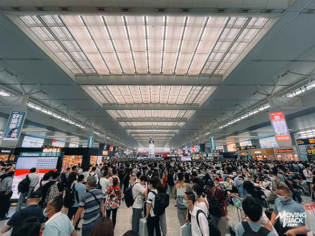 Crowded train station with people wearing masks, standing under a large, bright ceiling with signs and shops—reminiscent of the bustling energy found in Shanghai airports.