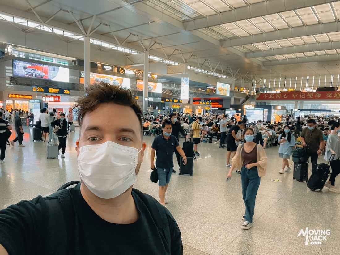 Man wearing a mask takes a selfie in a busy, modern train station—much like those found at Shanghai airports—with people walking and luggage around.