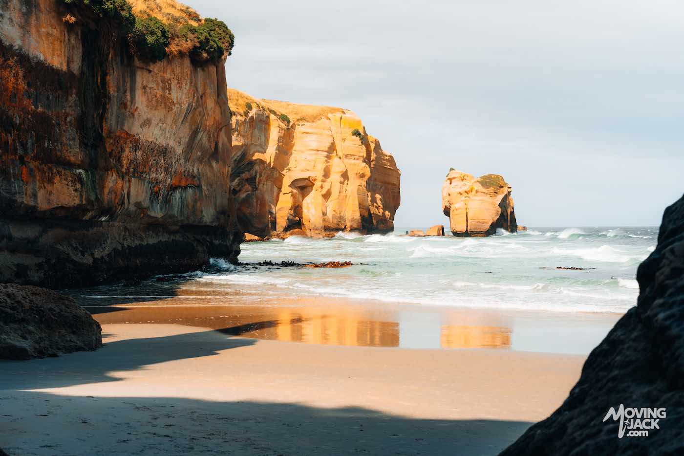 Een zandstrand met hoge, zonovergoten kliffen en rotsformaties die weerspiegeld worden in het ondiepe water onder een gedeeltelijk bewolkte hemel – een tafereel dat je doet afvragen: is Cathedral Caves een bezoek waard? Absoluut, vanwege de adembenemende schoonheid van de kust.
