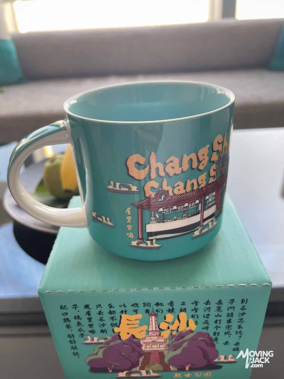Een turquoise mok met afbeeldingen van Changsha staat op een bijpassende doos, wat herinneringen oproept aan het zoeken naar een geschikte accommodatie in Changsha, met een knusse bank op de achtergrond.