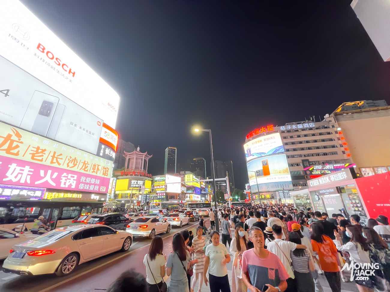 Een menigte mensen en auto's vult 's nachts een felverlichte stadsstraat vol reclameborden, wat de bruisende energie vastlegt die bezoekers doet afvragen waar ze in Changsha moeten verblijven.