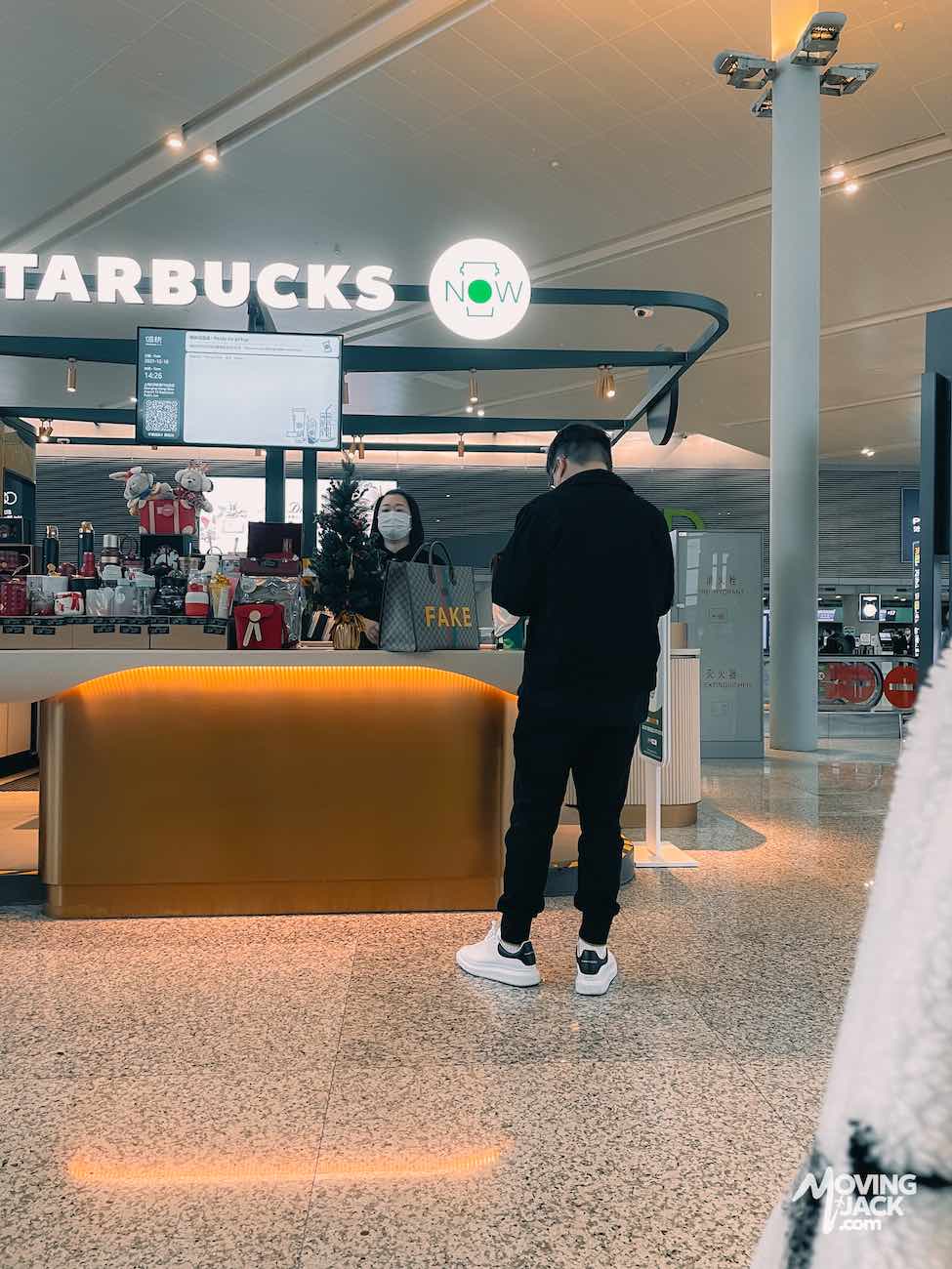 A man in black stands at a Starbucks counter in an airport in China; another person is behind the counter. A large bag with "FAKE" printed on it is visible.