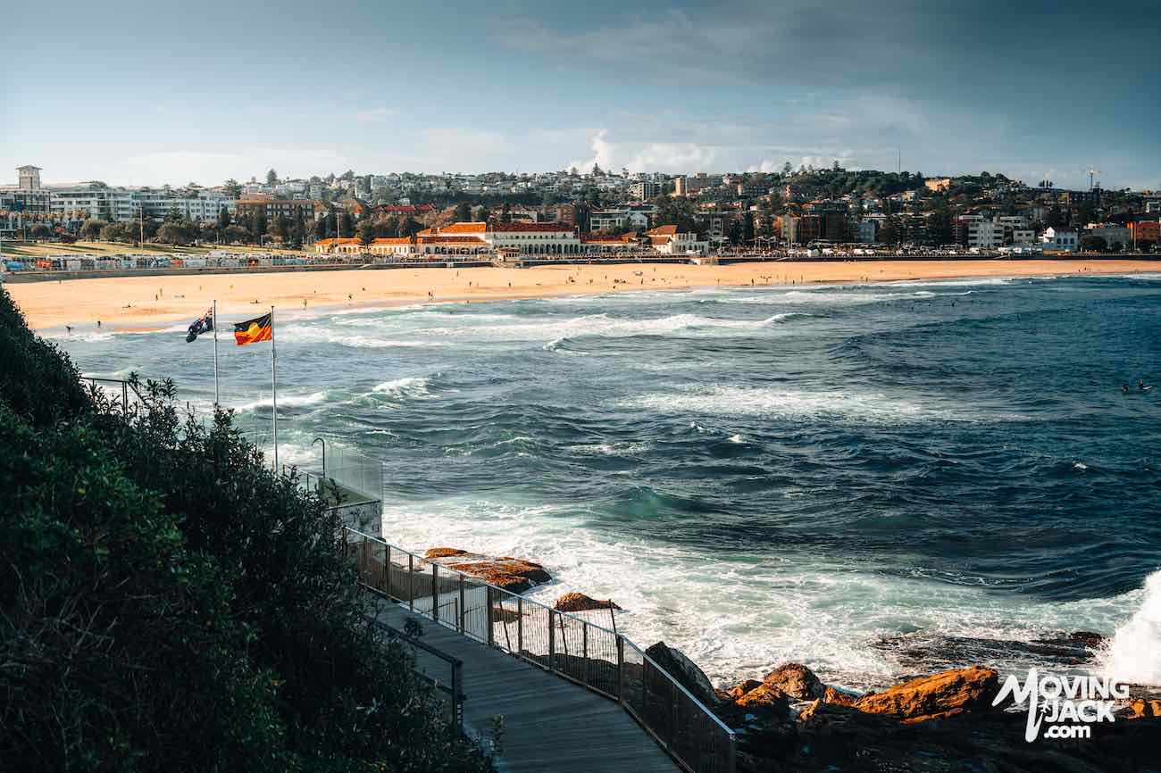 View of Bondi Beach with waves, sandy shore, distant buildings, and two flags near a wooden walkway