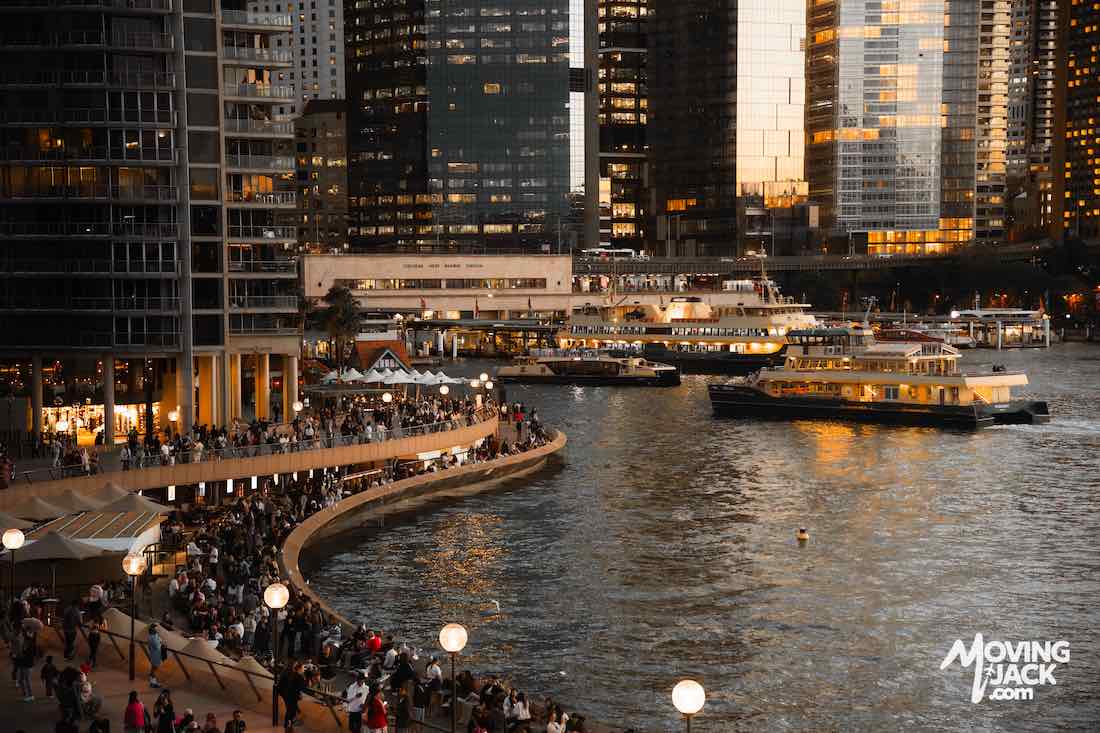 Crowds gather along a riverside promenade with ferries on the water and city skyscrapers at sunset.
