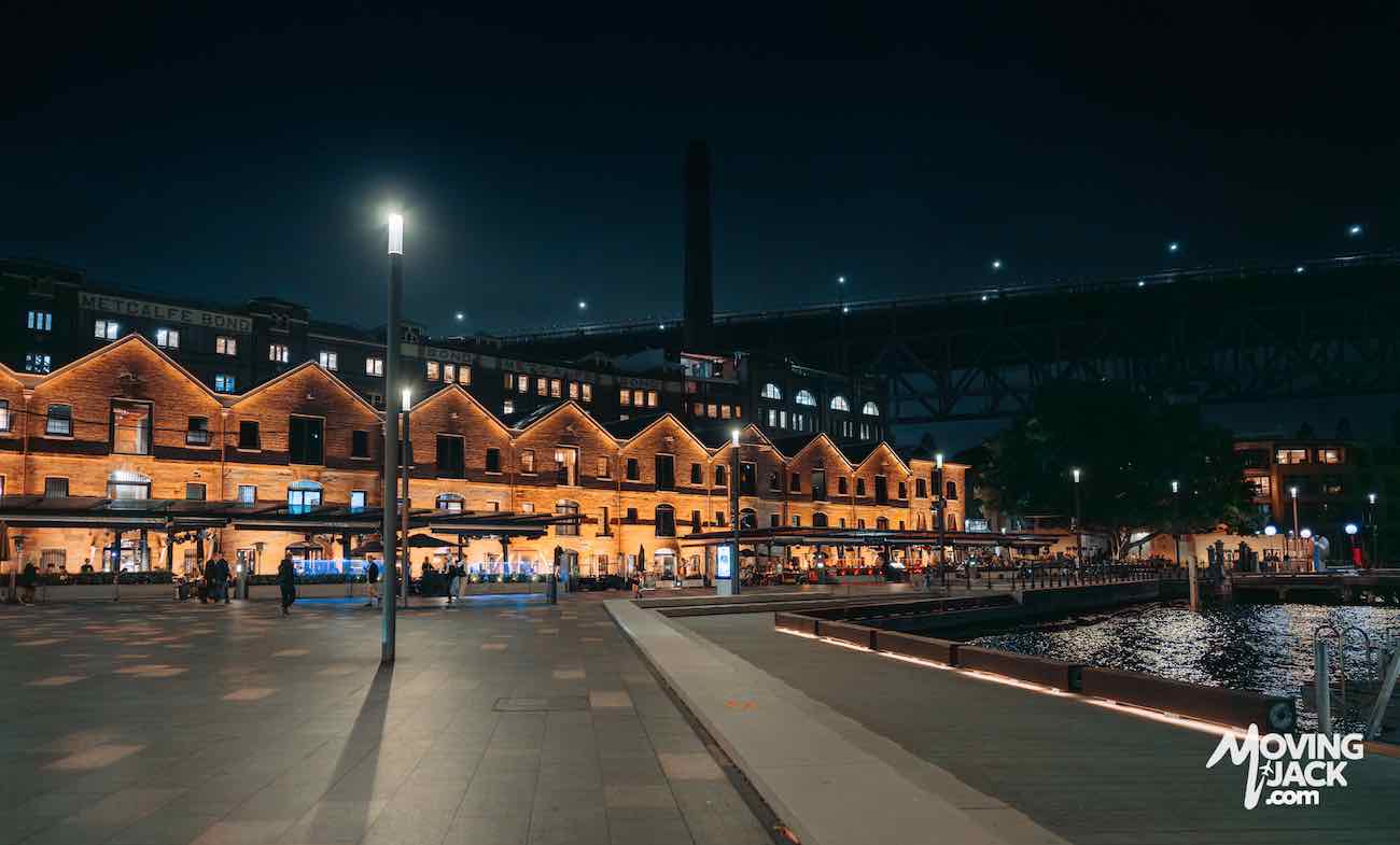 Night view of a waterfront promenade with lit historic buildings and people walking near the docks