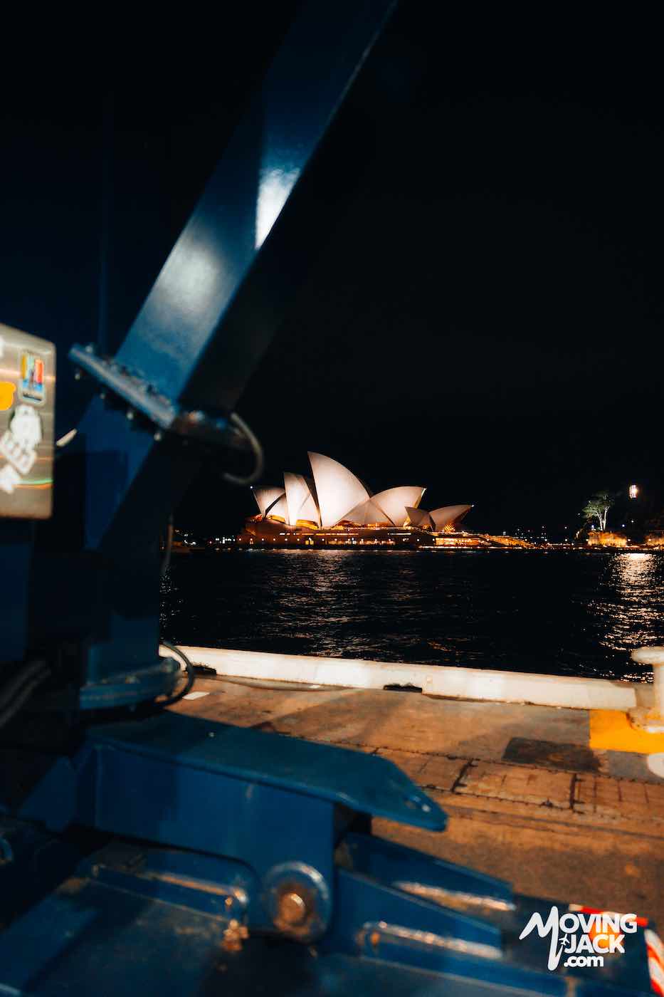 Sydney Opera House illuminated at night, viewed from a dock with machinery in the foreground.