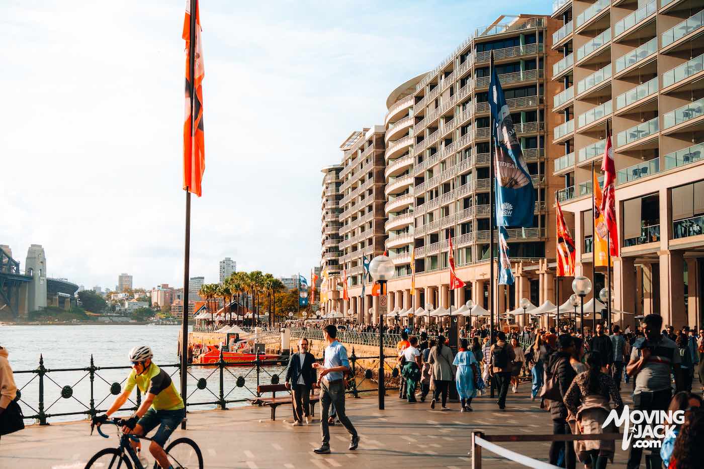 Crowd walking along a waterfront promenade lined with flags and modern buildings.