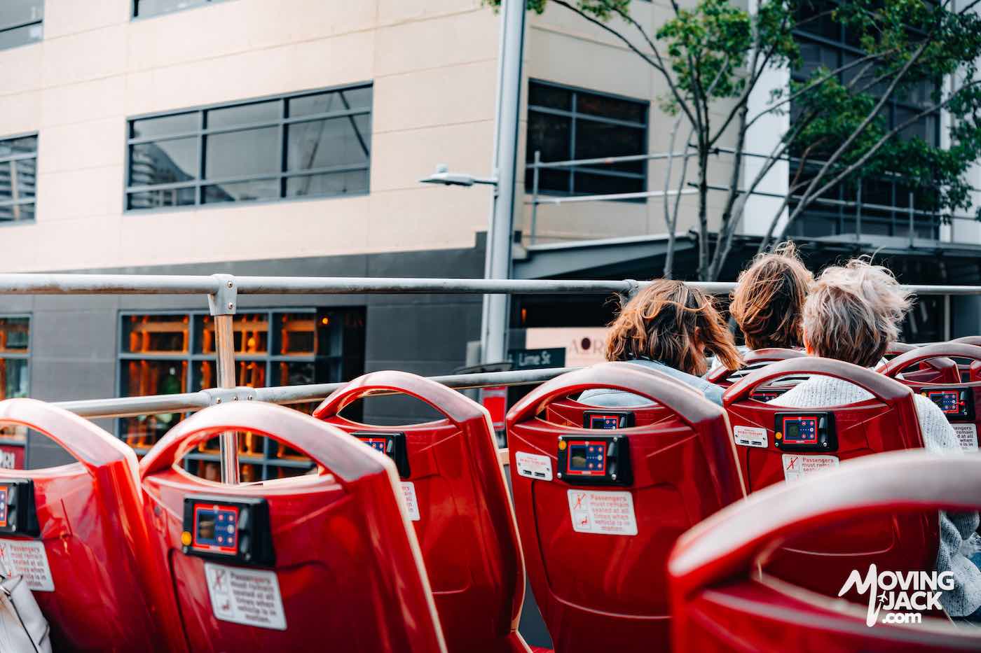 Three people sit on the upper deck of a red open-top tour bus passing by city buildings and trees