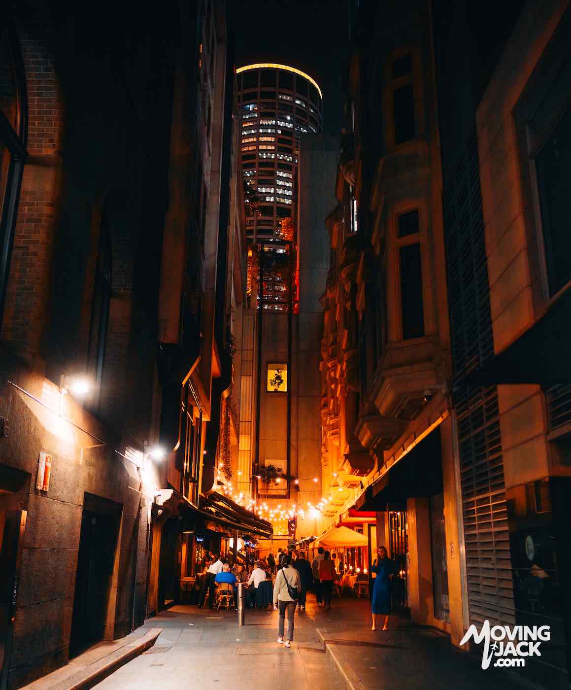 A narrow city alley at night, warmly lit, with people walking and a tall modern building in the background.