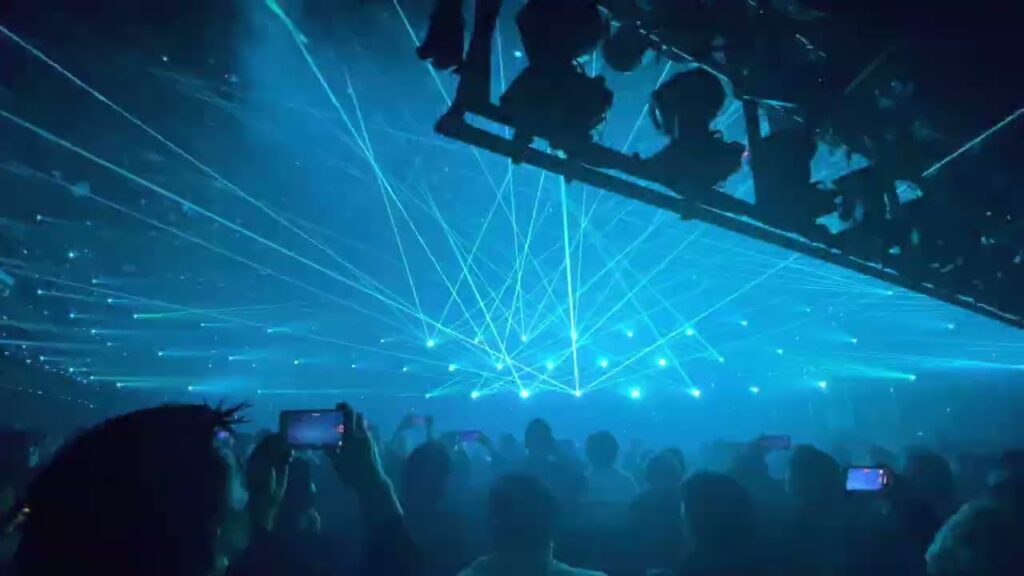 A crowd reminiscent of a lively company meeting watches the concert as vibrant blue and green laser lights illuminate the stage.