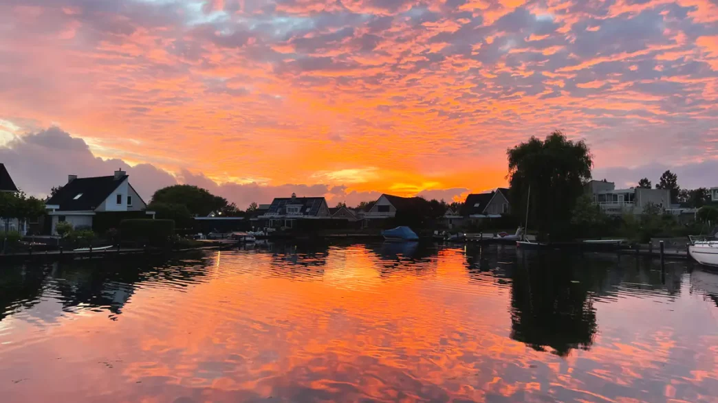 De zonsondergang boven een kalm meer met silhouetten van huizen en bomen weerspiegelt feloranje en roze wolken, en legt zo de prachtige lucht boven Nederland vast.