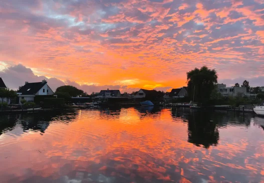 De zonsondergang boven een kalm meer met silhouetten van huizen en bomen weerspiegelt feloranje en roze wolken, en legt zo de prachtige lucht boven Nederland vast.